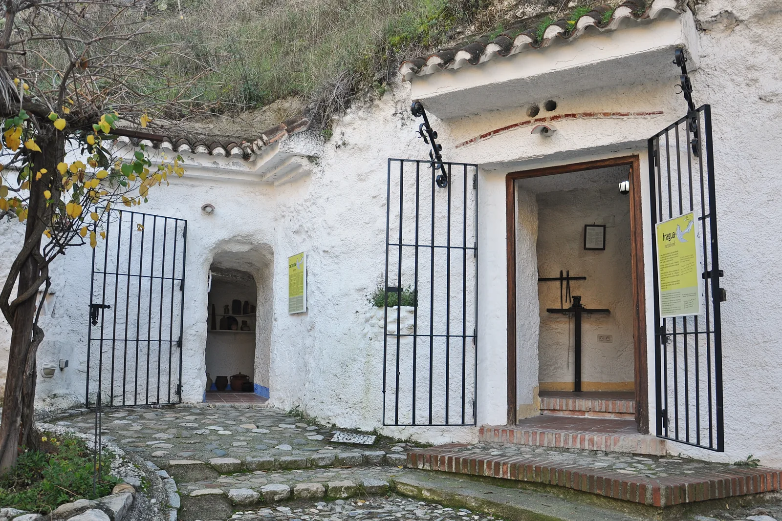 Sacromonte Mağaraları Müzesi'ndeki bir mağara binasının badanalı ön cephesi; kiremitli çatı kenarı, demir kapılar ve bir limon ağacı